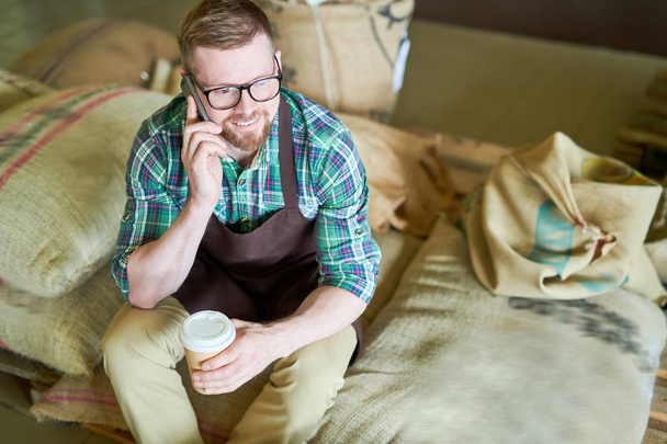 Vysoký úhel portrét pohledný mladý muž, sedící na hromadu pytlovina pytle držení papíru šálek kávy a mluvit po telefonu, při přijetí přerušení v klasickem pražírny, zkopírujte prostor - Fotografie, Obrázek