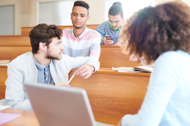 Sério pensivo bonito estudante hispânico menino em camisa quadriculada ouvir colega de grupo enquanto eles conversam durante a pausa na sala de aula
 - Foto, Imagem