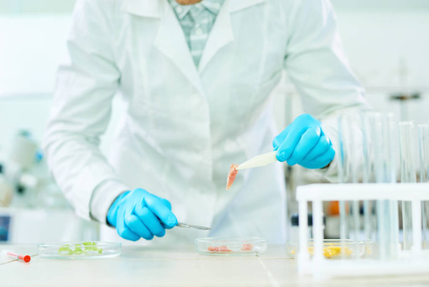 Close-up shot of unrecognizable researcher wearing white coat and rubber gloves standing at laboratory bench and studying sample of artificial meat with help of scalpel. - Zdjęcie, obraz