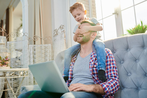 Portrait of cute little boy covering eyes of handsome dad using laptop while sitting on his shoulders and playing at home in sunlight - Photo, Image