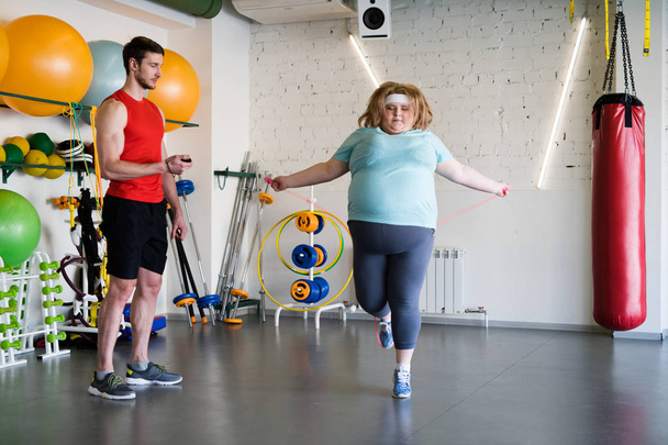Full length portrait of big woman jumping with rope in gym while training with fitness instructor - Valokuva, kuva