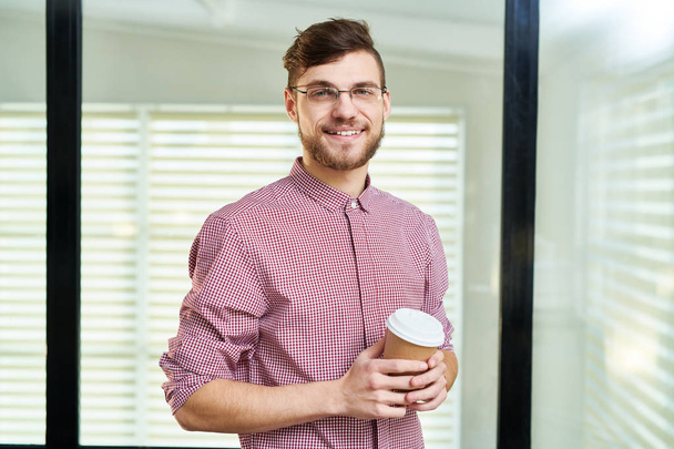 Retrato en la cintura de un guapo oficinista barbudo en vasos sosteniendo una taza de café y sonriendo a la cámara
 - Foto, Imagen