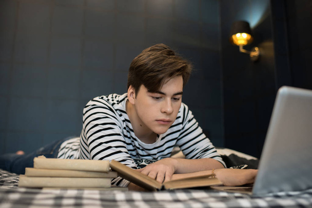 Ελκυστικό νεαρό αρσενικό ανάγνωση σωρός από βιβλία και χρήση φορητού υπολογιστή ενώ ξαπλωμένος στο κρεβάτι. - Φωτογραφία, εικόνα