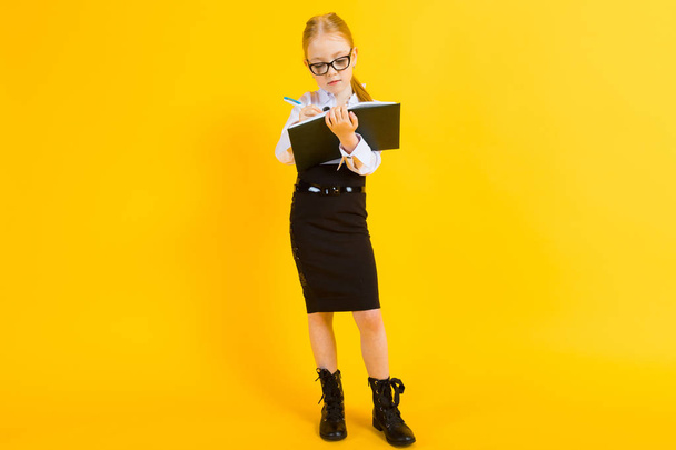 Girl with red hair on a yellow background. A charming girl in transparent glasses writes a pen in her notebook. - Photo, Image