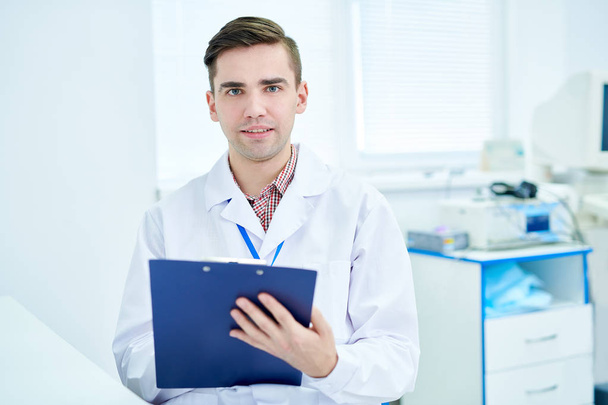 Waist up portrait of young doctor holding clipboard and looking at camera posing in modern office, copy space - Fotografie, Obrázek