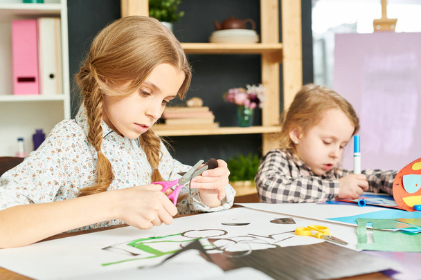 Pretty girl with two braids cutting shape from colored paper while her little sister wrapped up in drawing with felt-tip pens, interior of cozy living room on background - Фото, изображение