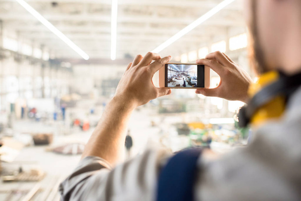 Over shoulder view of unrecognizable inspector wearing ear protectors standing at spacious production department of modern plant and taking picture on smartphone while carrying out inspection. - Foto, imagen