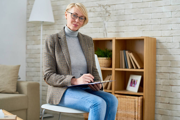 Porträt einer blonden Psychiaterin, die im Büro mit Klemmbrett posiert und in die Kamera schaut, Kopierraum - Foto, Bild