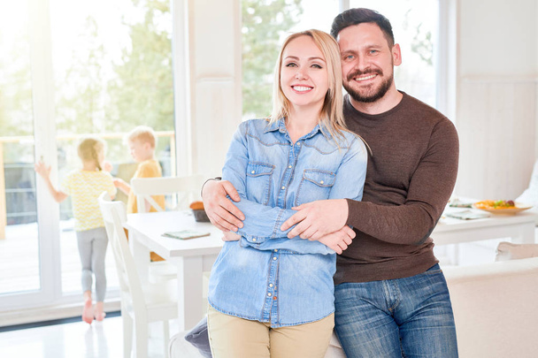 Portrét šťastné milující pár pózuje při pohledu na fotoaparát v moderním obývacím pokoji se dvě děti si hrají v pozadí, kopírování prostor - Fotografie, Obrázek