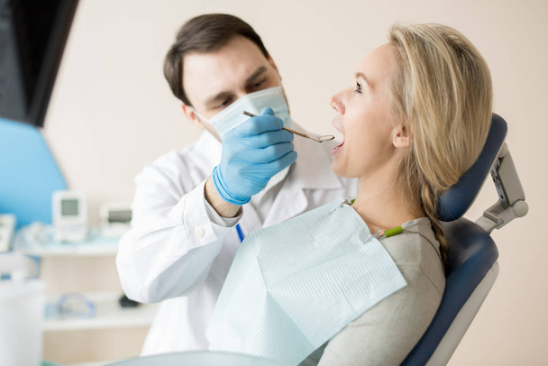 Вид сбоку на девочку, сидящую в кресле, пока стоматолог с маленьким зеркалом изучает состояние зубов
.  - Фото, изображение