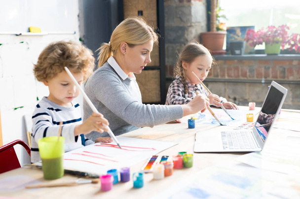 Jonge leraar helpen haar leerlingen kunnen een tekening met aquarel verven aan de grote tafel in art studio - Foto, afbeelding