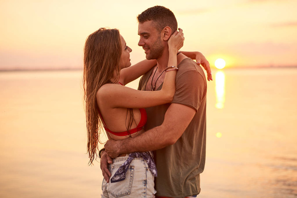 Onnellinen hellä kaunis nuori pari rento vaatteet halaavat toisiaan ja katsellen silmiin aikana päivämäärä merellä auringonlasku
 - Valokuva, kuva
