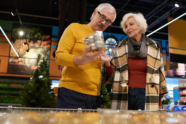 Retrato de alegre pareja de personas mayores comprando en el supermercado eligiendo decoraciones navideñas
 - Foto, Imagen