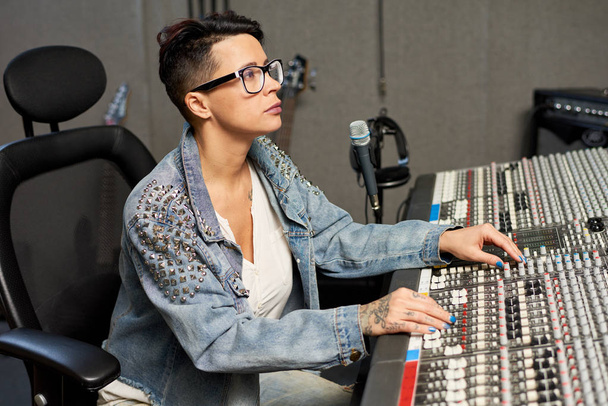 Вид сбоку стильной женщины в джинсах, использующей пульт управления во время работы в звуковой студии
.  - Фото, изображение
