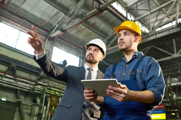 hüfthoch Porträt eines gutaussehenden Geschäftsmannes, der nach oben zeigt, während er etwas mit einem Fabrikarbeiter diskutiert, der ein digitales Tablet in der Hand hält, Kopierraum - Foto, Bild