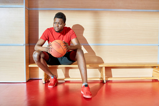Αφρικανικός-αμερικανικός αθλητικός τύπος κρατώντας μπάλα μπάσκετ και αναζητούν μακριά ενώ ξεκουραζόταν σε πάγκο στο γυμναστήριο.  - Φωτογραφία, εικόνα