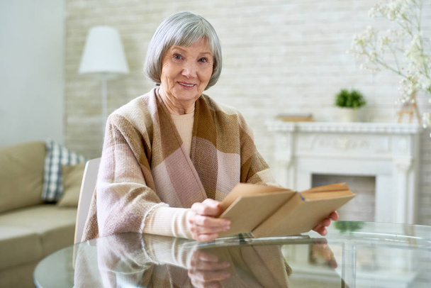 Portret van aardige senior dame gehuld in geruite sjaal camera kijken en zachtjes lacht tijdens het lezen van boek zit op glazen tafel thuis, kopie ruimte - Foto, afbeelding