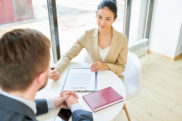 High angle portrait of pretty young businesswoman smiling and holding contract looking at client discussing terms of beneficial deal during meeting at table by window in modern office, copy space - Photo, Image