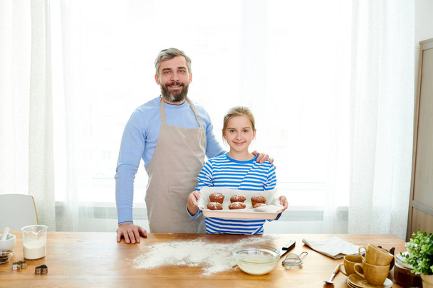Portret van knappe volwassen man poseren met meisje houden bakplaat met trots presenteren muffins permanent in moderne keuken thuis - Foto, afbeelding