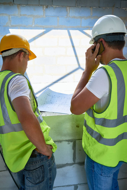 Vissza view portréja két építőmunkások visel hardhats belül befejezetlen épület emelet tervek megvitatása - Fotó, kép