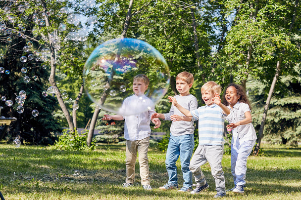 Многонациональная группа детей, веселящихся с большими мыльными пузырями, проводит теплый летний день в общественном парке, освещенном солнечными лучами
 - Фото, изображение