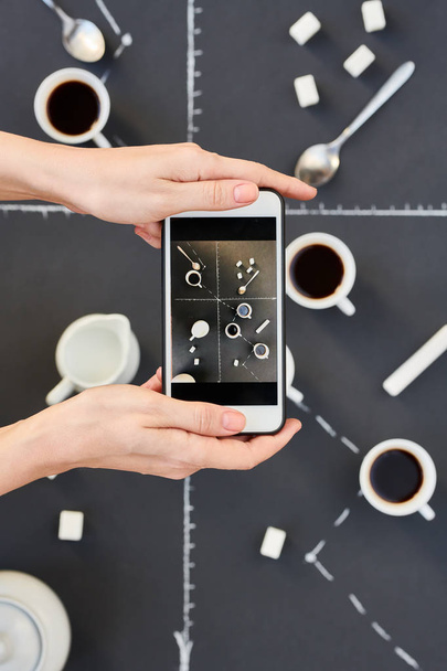 Above  view of female hands taking smartphone photo of coffee cups and sugar cubes laid over  grey background with chalk graph on it , business, statistics, development or deadline concept, copy space - 写真・画像