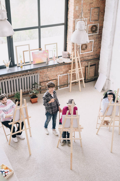 Portrait von Studenten beim Malen an Staffeleien im Kunstatelier sitzend mit einer Kunsterzieherin, die sie in einem großzügigen, sonnenbeschienenen Loftraum beobachtet - Foto, Bild