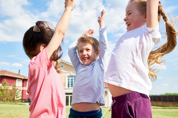 Retrato de la cintura hacia arriba de tres niños emocionados animando felizmente y levantando las manos mientras se divierten al aire libre, jugando en el césped verde en el patio delantero
 - Foto, Imagen