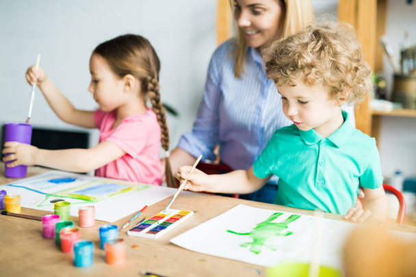 Μικρό αγόρι με πινέλο σχεδίασης του εικόνα με χρώματα μαζί με την αδελφή και μητέρα βοηθώντας τους - Φωτογραφία, εικόνα