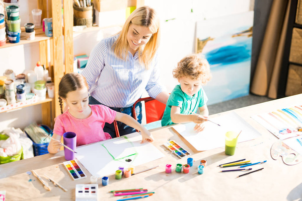 Vue en grand angle de deux enfants peignant des tableaux en classe d'art avec un jeune professeur qui les aide
 - Photo, image