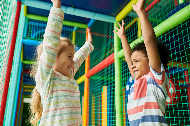 Colorful waist up portrait of happy boy and girl facing each other and raising hands in excitement while having fun in children play area - Fotoğraf, Görsel
