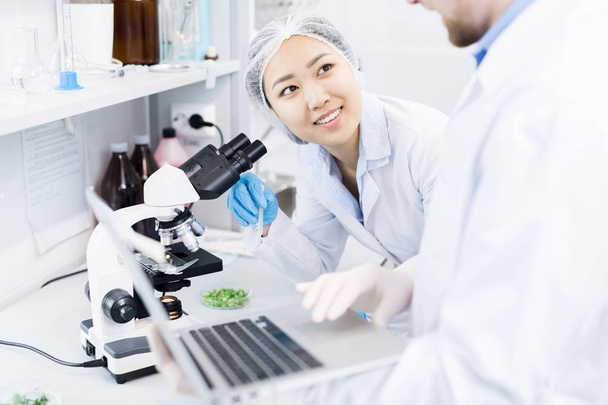 Crop view of smiling female microbiologist standing at eyepiece of microscope with samples of green vegetables looking at colleague typing results of analysis on laptop - Photo, Image