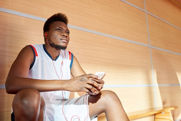 Handsome black man in basketball uniform sitting on bench and enjoying nice music while resting during training.  - Photo, Image