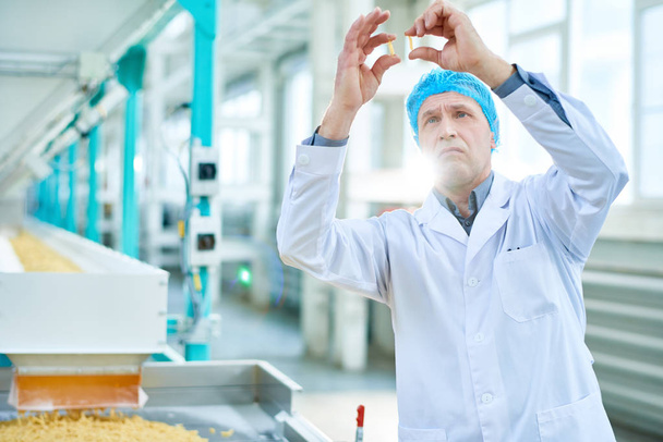 Waist up portrait of senior factory worker doing quality inspection in food industry holding two macaronis standing by conveyor belt, copy space - Photo, Image