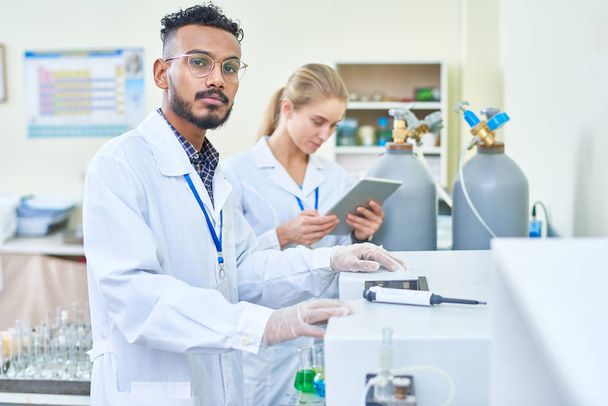 Serious handsome young Arabian scientist in lab coat choosing program for machine while working in laboratory, his female assistant using tablet behind him - Foto, immagini