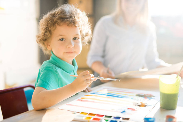 warmgetöntes Porträt des niedlichen kleinen Jungen, der in die Kamera schaut, während er Bilder malt, die im hellen Sonnenlicht am Tisch sitzen - Foto, Bild