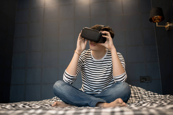 Giovane maschio utilizzando auricolare realtà virtuale mentre seduto sul letto in camera oscura
. - Foto, immagini