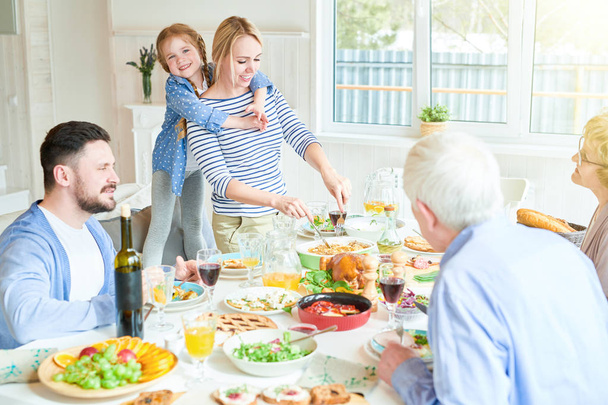 Portrait of happy family enjoying dinner sitting round festive table with delicious dishes, cute redhead girl hugging mom serving food during holiday celebration in modern sunlit apartment, copy space - Photo, Image