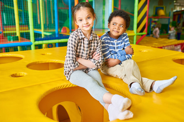 Full length portrait of cute girl and African-American boy posing sitting on top of cheese like box while having fun in large indoor play area for children - Foto, Imagem