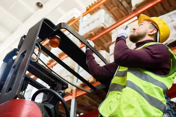 Low angle portrait of young man sitting in forklift and using walkie-talkie while moving goods in warehouse, copy space - Fotografie, Obrázek