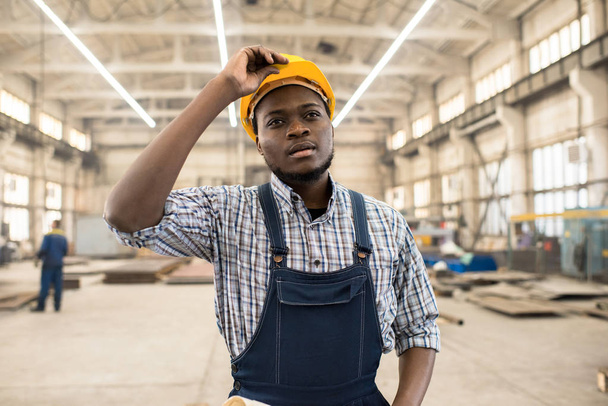 Portrait shot of young African American technician wearing overall and protective helmet standing at spacious production department of modern plant and controlling manufacturing process - Photo, Image