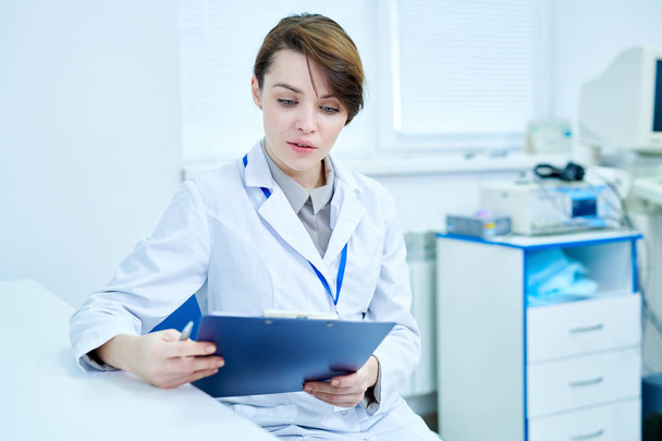 Médecin femme en manteau blanc assis au bureau tenant le presse-papiers et lisant attentivement
. - Photo, image