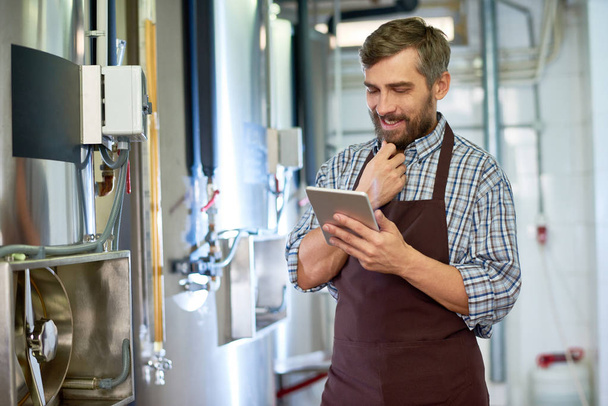Портрет усміхненого бородатого техніка, який носить перевірену сорочку та фартух за допомогою цифрового планшета, під час налаштування обладнання на сучасній пивоварні
 - Фото, зображення