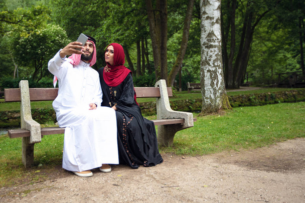 Young beautiful Arabian couple casual and hijab, Abaya, taking a selfie on the lawn in summer park. - 写真・画像