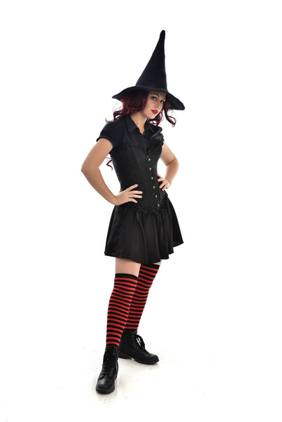 Portret van de volledige lengte van red haired meisje dragen zwarte heks kostuum en puntige hoed. staande pose, geïsoleerd op witte studio achtergrond. - Foto, afbeelding