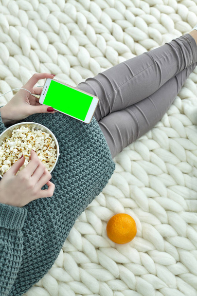 Очень уютная картина для осени, сентября, октября, ноября. Девушка смотрит кино по телефону в наушниках и ест попкорн. Зеленый экран на мобильном телефоне для изображения, текста, информации
. - Фото, изображение