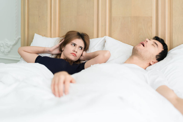 Zirytowany Asian żona blokuje jej uszy od hałasu chrapanie w sypialni w domu męża. Młoda para ma problem z chrapaniem mężczyzny. - Zdjęcie, obraz