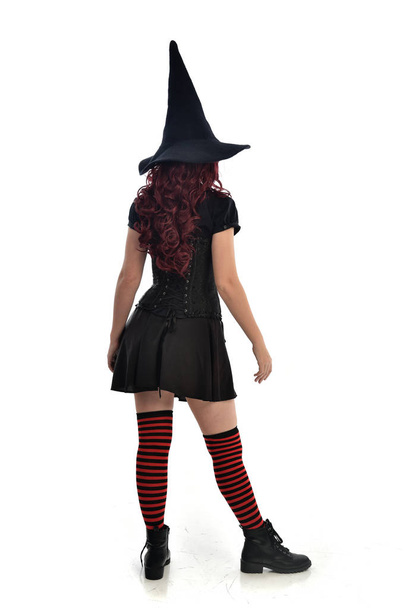 retrato de comprimento total da menina de cabelos vermelhos vestindo traje de bruxa preta e chapéu pontiagudo. postura de pé com as costas para a câmera, isolado no fundo do estúdio branco
. - Foto, Imagem