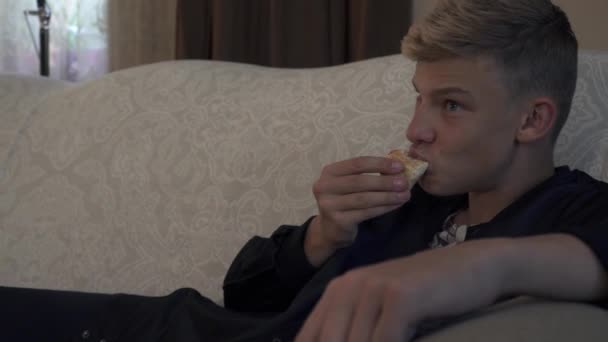 Young man eats pizza laying at sofa - Felvétel, videó