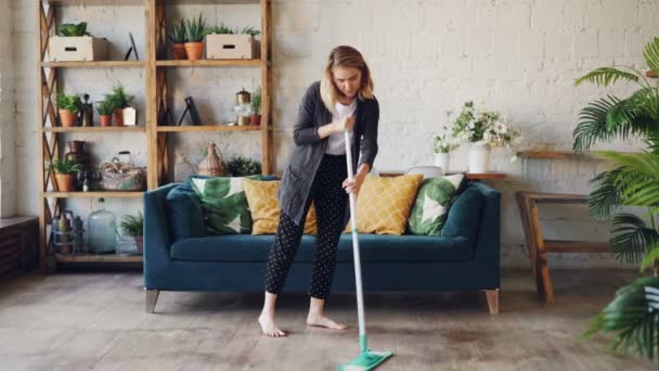 Pretty blond housewife is doing housework mopping wooden floor in beautiful flat cleaning under sofa. Girl is concentrated on her occupation, she is barefoot. - Кадры, видео